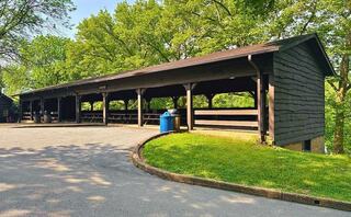 A wooden pavilion sits along a winding path, surrounded by lush greenery, offering shaded seating and a nearby trash bin.