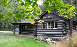 A rustic log cabin with the sign "Camp Russel" surrounded by lush greenery and trees, offering a serene natural setting.
