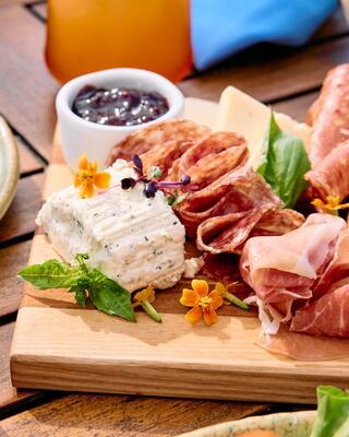A wooden platter features assorted meats, cheese, herbs, and edible flowers, creating a vibrant and appetizing presentation.
