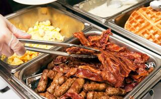 A hand uses tongs to serve crispy bacon from a buffet, alongside sausage links and scrambled eggs in metal trays. Waffles are present too.