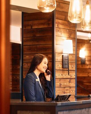 A smiling receptionist answers a phone at a stylish front desk, surrounded by warm wooden paneling and modern pendant lights.