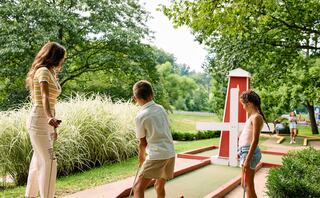 A woman supervises two children playing mini-golf in a lush green park, surrounded by trees and a sunny atmosphere.