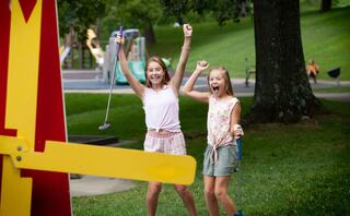 Two excited girls celebrate joyfully at a park, with playground equipment in the background. Their energy and happiness shine through.