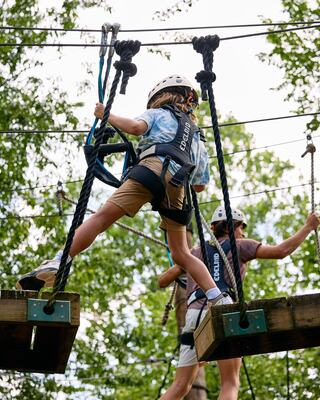 A child navigates an aerial adventure course, balancing on ropes with a harness while another participant looks on from a nearby platform.