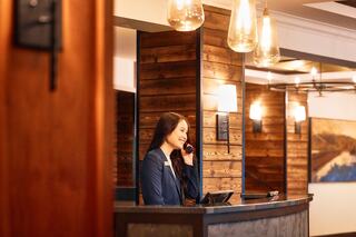 A smiling receptionist speaks on the phone at a chic, wooden-paneled front desk with warm lighting and modern decor.