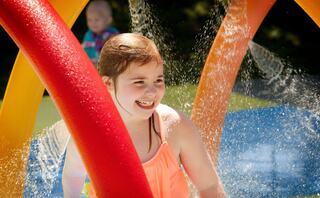 A smiling girl in a vibrant swimsuit plays joyfully under colorful water arcs, enjoying a sunny day at a splash park.