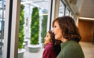 A woman and a girl admire greenery through a glass window, sharing a moment of connection and curiosity in a bright, modern space.